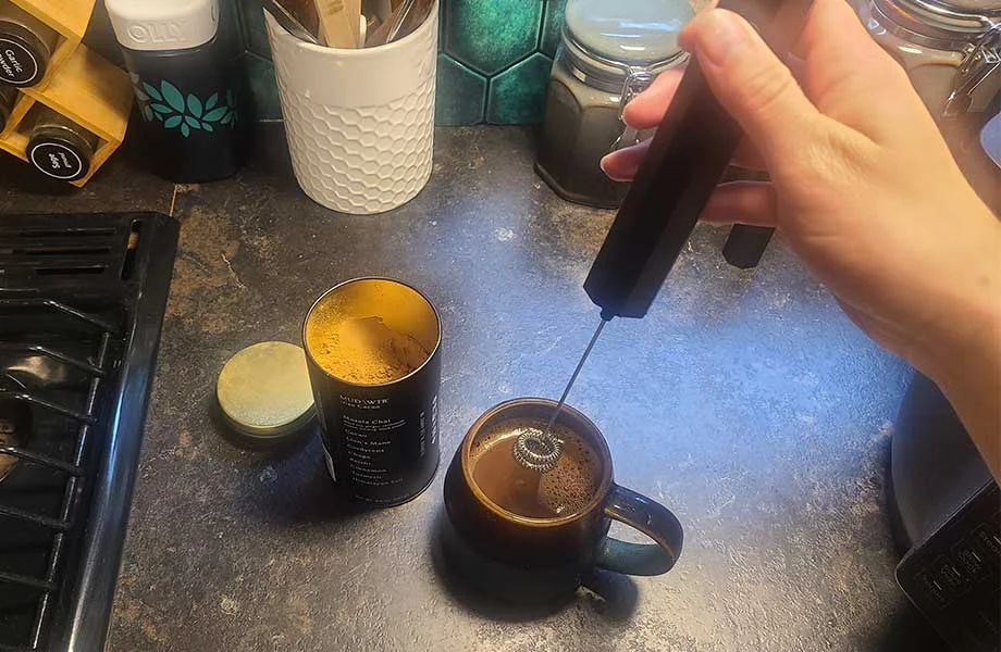 Using an electric hand frother to mix the mushroom coffee in a mug.