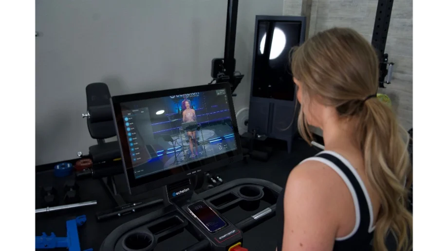 A woman using a treadmill with a large integrated touchscreen display showing a fitness class.