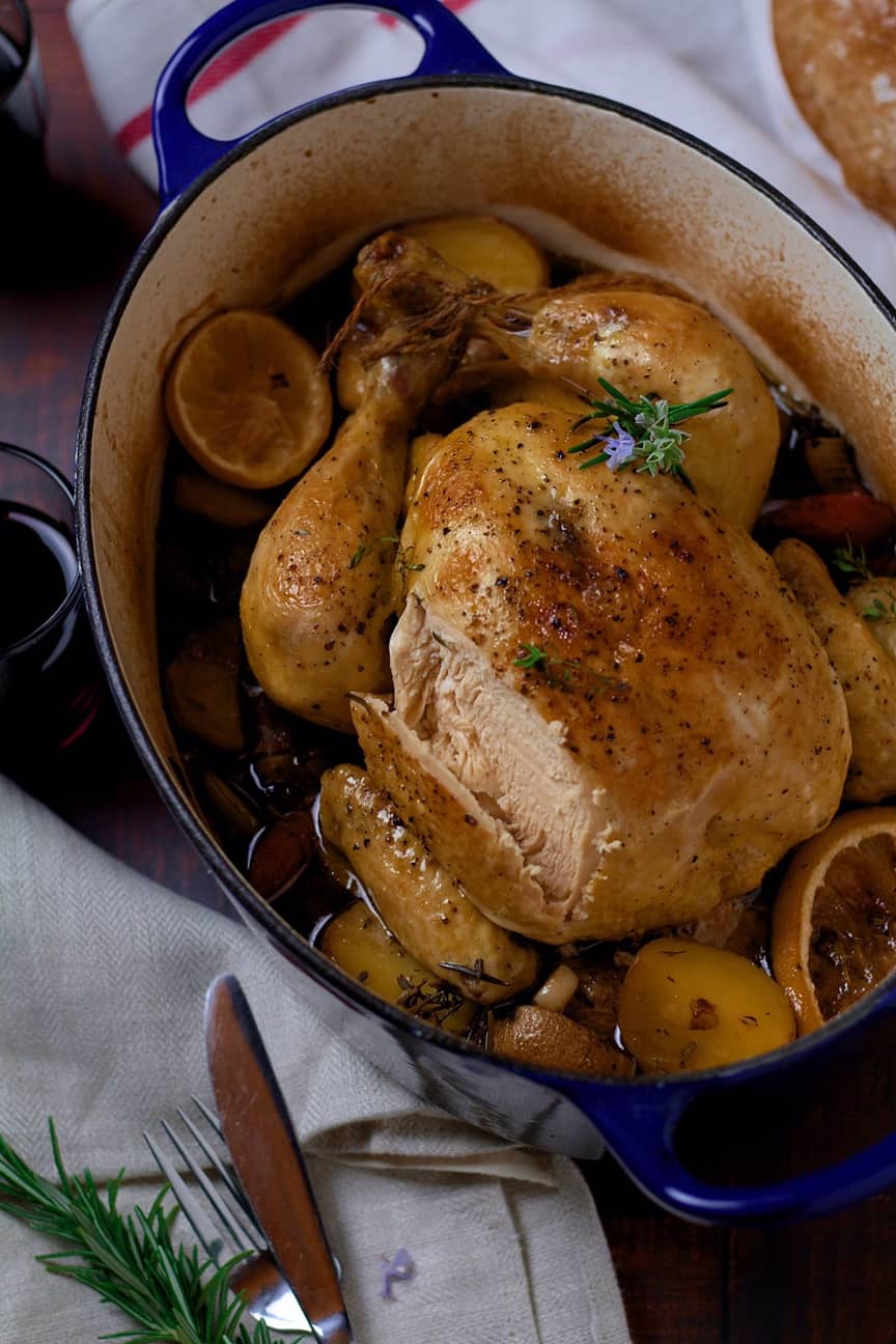 A whole roasted chicken with potatoes and lemon slices inside a blue enamel Dutch oven.
