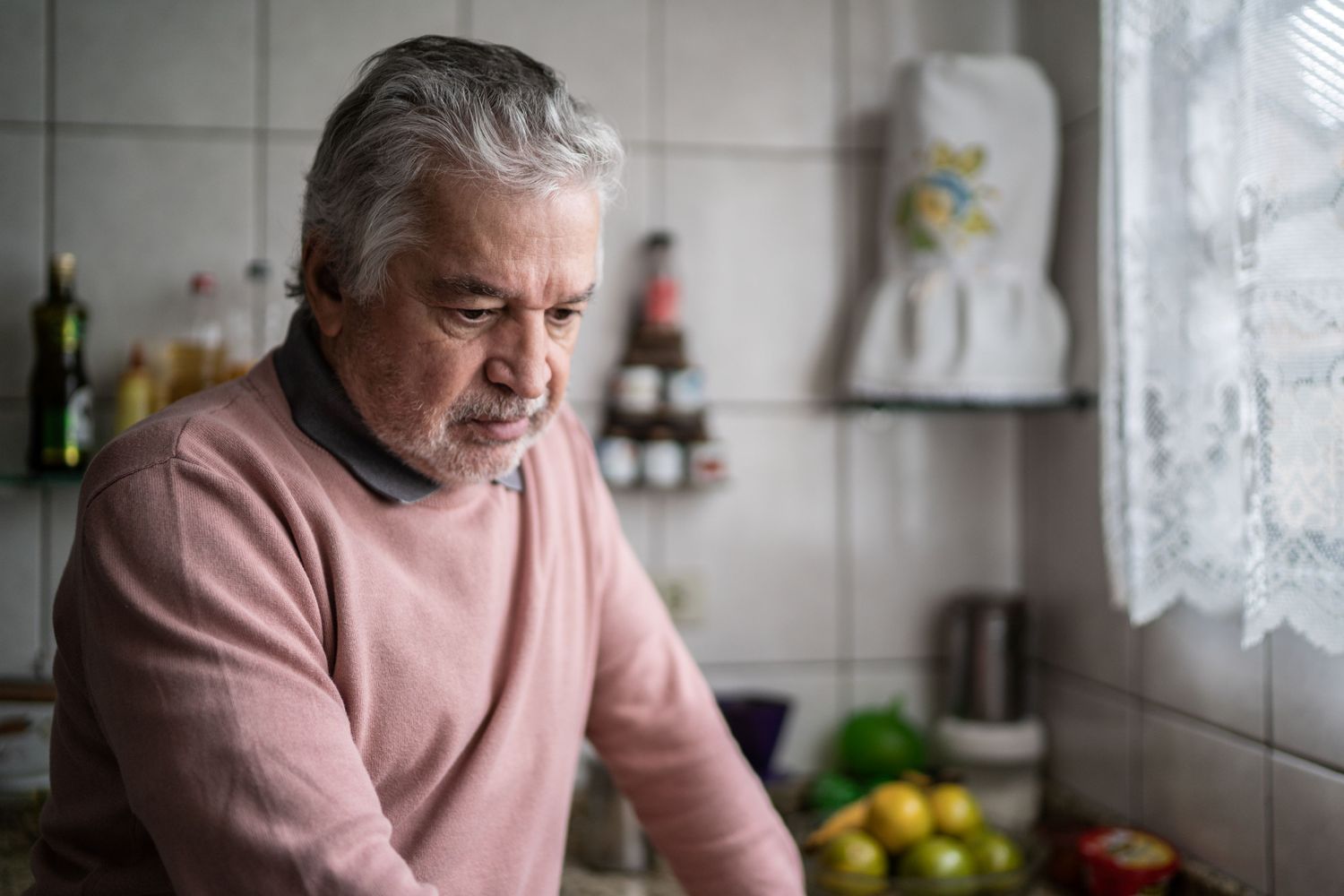 A person with gray hair standing in a kitchen environment, looking reflective.