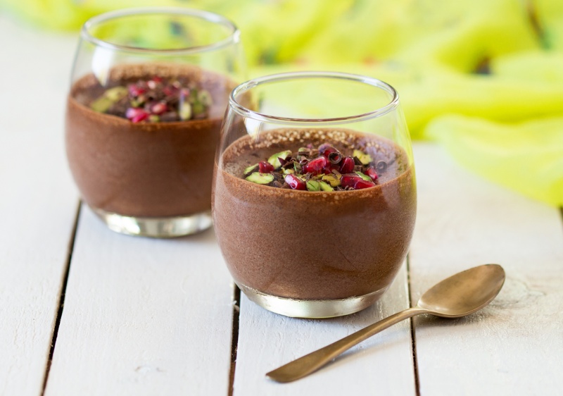 Two glasses of dark chocolate mousse garnished with bright red pomegranate seeds and crushed green pistachios.