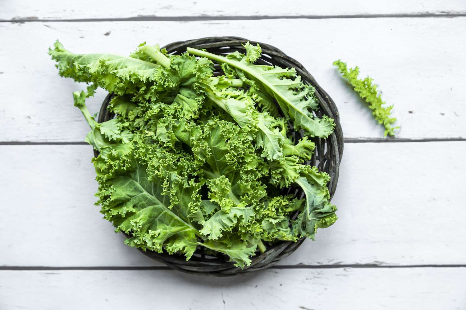 A woven basket filled with fresh green kale on a white wooden surface.