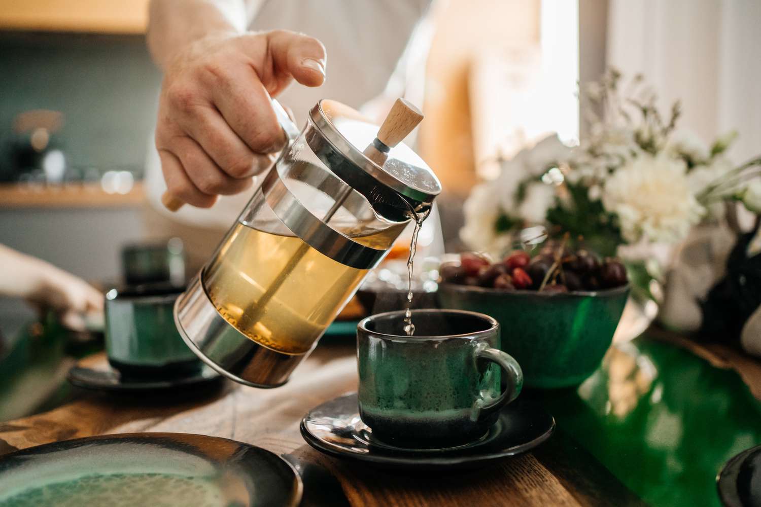 A person pouring warm tea from a glass press into a ceramic mug on a wooden table.