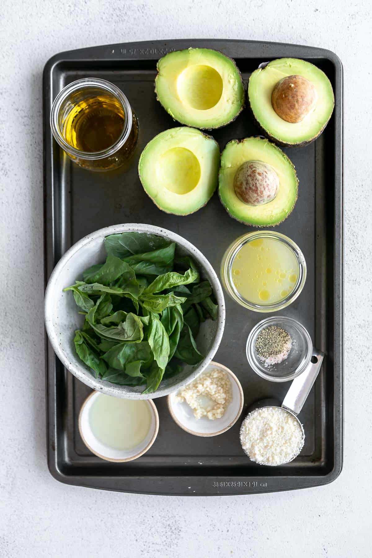 Flat lay of raw ingredients including ripe avocados, fresh basil leaves, cashews, and lemons in small bowls.
