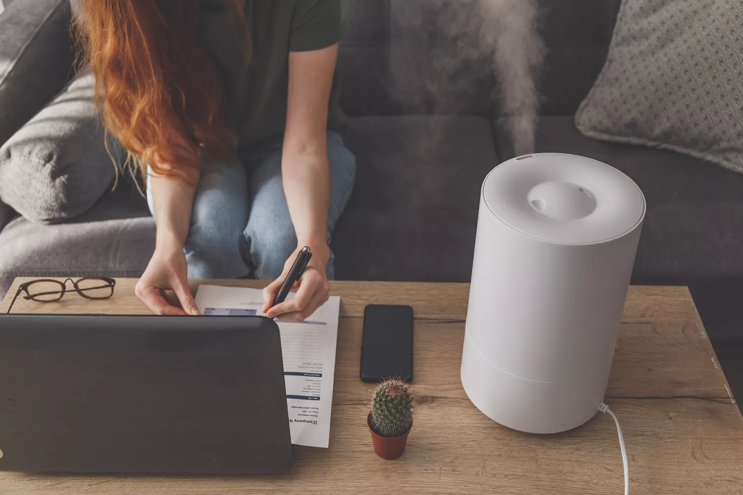 A woman working on a laptop at a desk with a white humidifier emitting a steady mist nearby.
