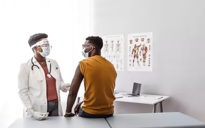 A masked healthcare provider consulting with a patient in a clinical exam room with anatomical charts.