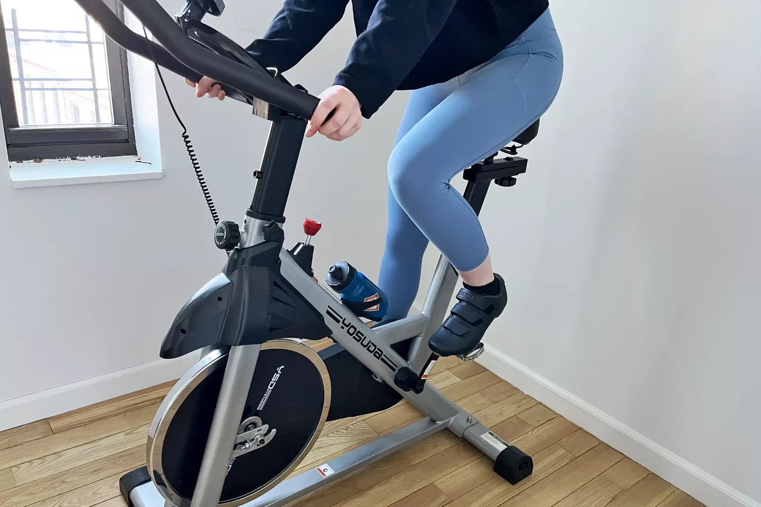 A person riding a YOSUDA indoor cycling bike in a bright room with hardwood floors.