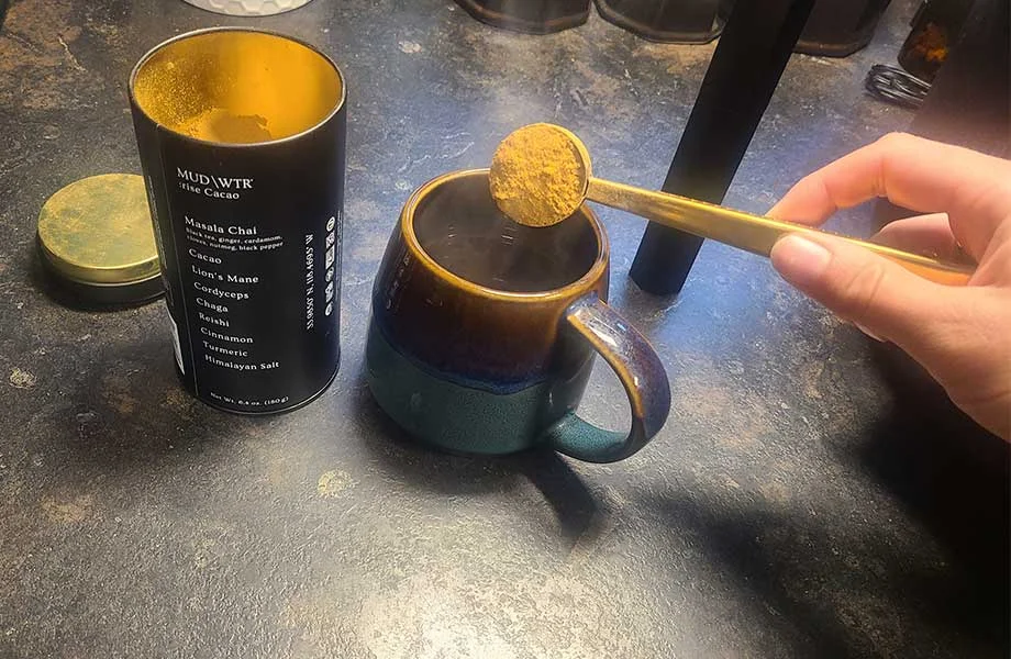 A wooden scoop lifting MUD\WTR powder into a white ceramic mug.