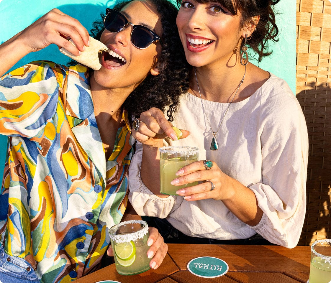 A group of friends laughing and sharing non-alcoholic drinks and appetizers at an outdoor patio table.