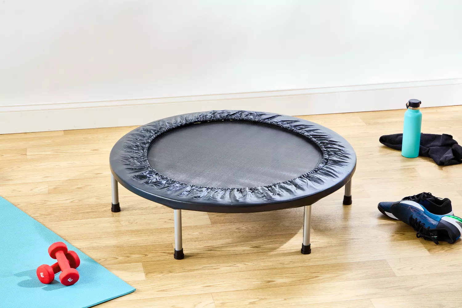 A mini trampoline on a wooden floor surrounded by dumbbells and a water bottle.