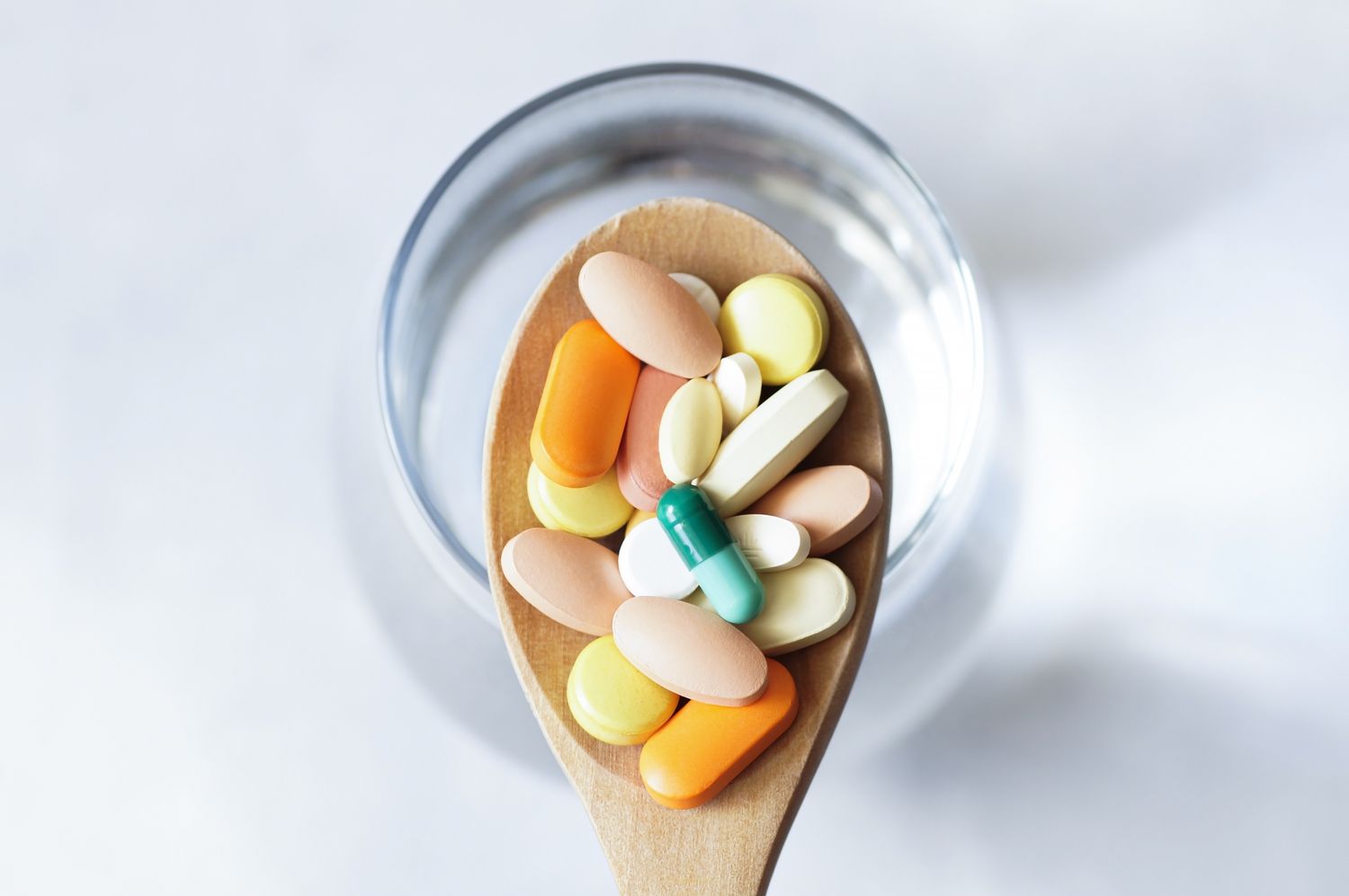 A wooden spoon holding various medical capsules and tablets next to a glass of water.