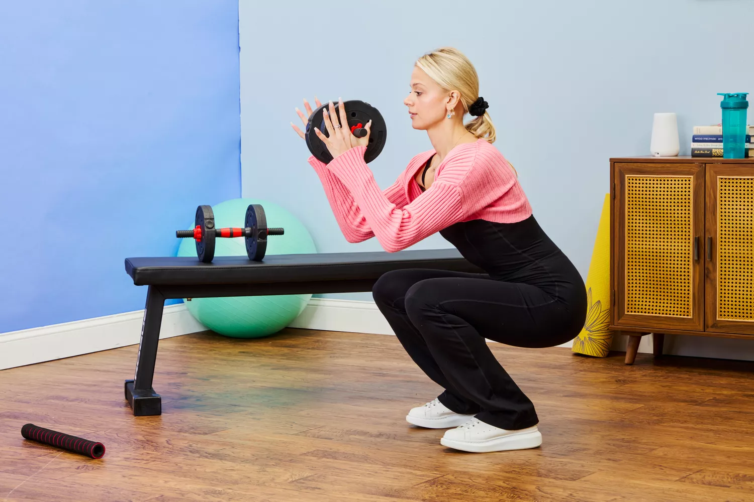 A woman performing squats with a weight plate in a home gym containing a bench and exercise ball.