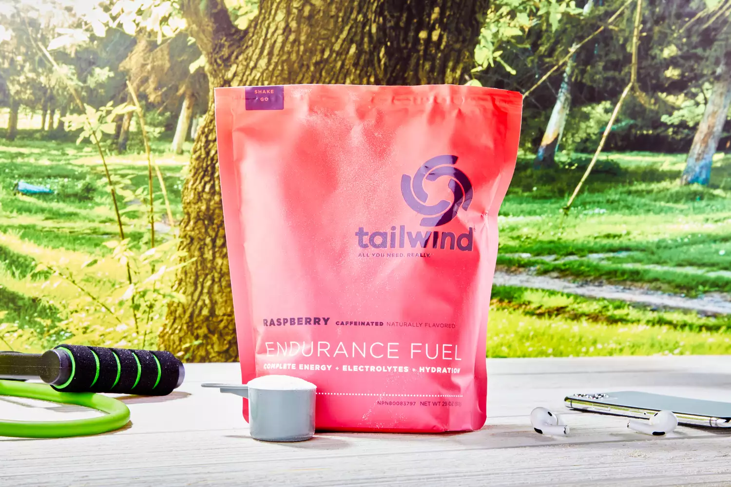 A pink bag of Tailwind Raspberry Caffeinated Endurance Fuel on a table with a jump rope and earbuds in an outdoor park setting.