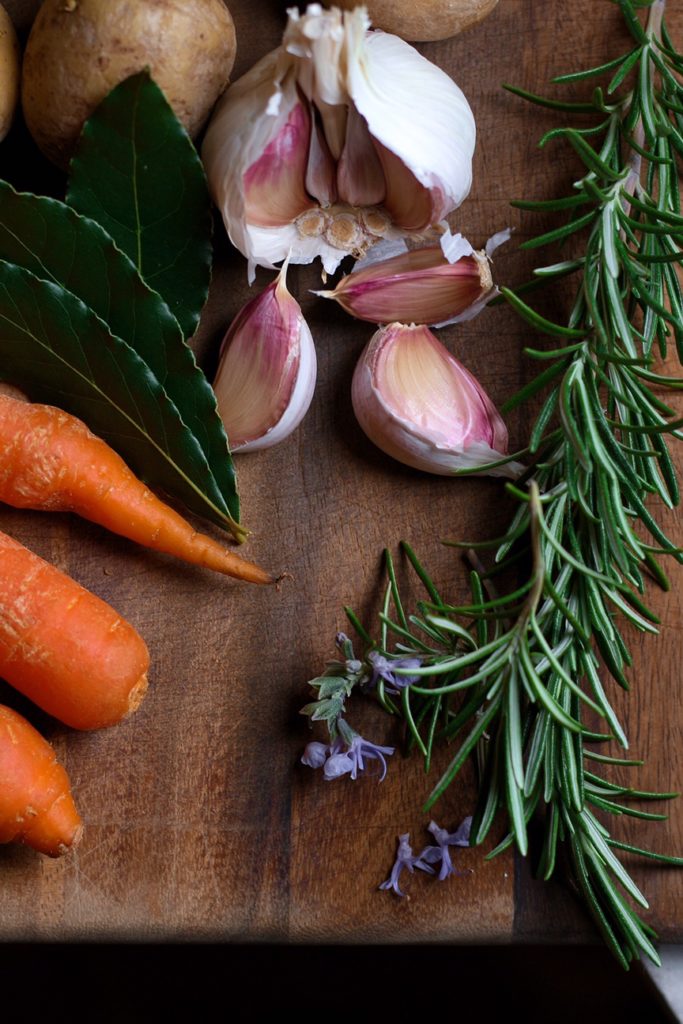 Chopped carrots, potatoes, garlic cloves, and sprigs of fresh rosemary on a wooden cutting board.