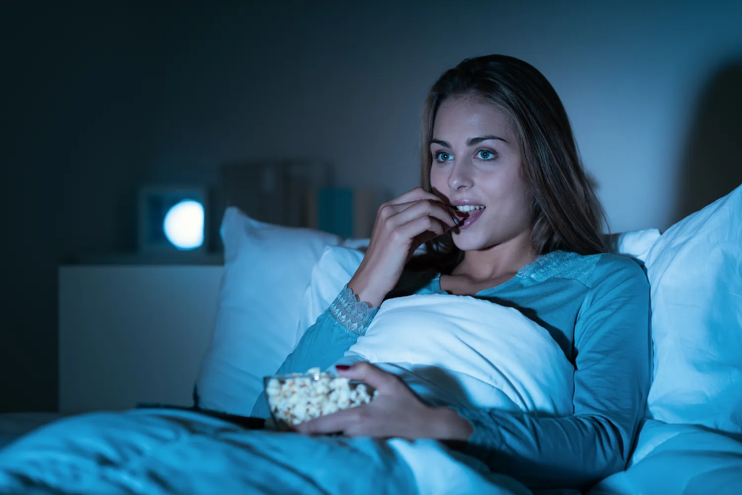 A person sitting in bed at night watching television and eating popcorn.