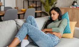A woman sitting on a sofa holding her stomach in visible discomfort.