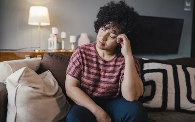 A woman holding her head in discomfort while sitting on a couch at home.