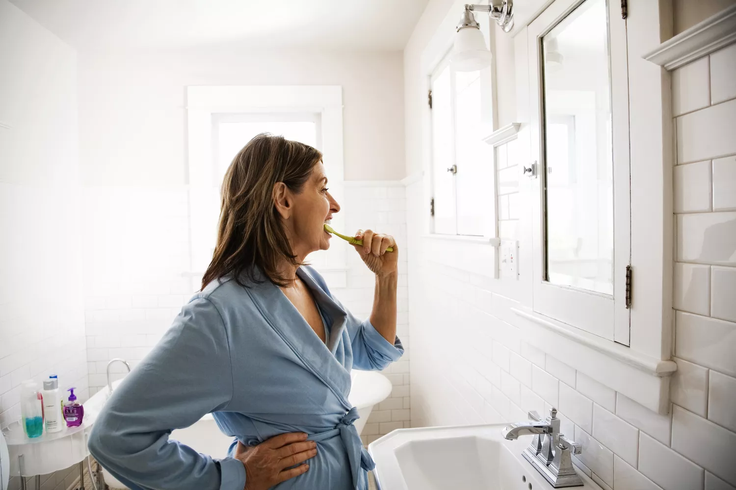 A woman appearing visibly tired and weary while brushing her teeth in a bathroom.
