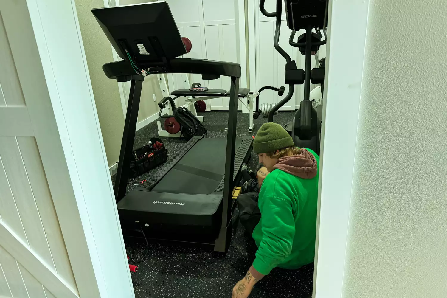 A technician kneeling beside a NordicTrack treadmill inspecting the motor base and belt alignment.