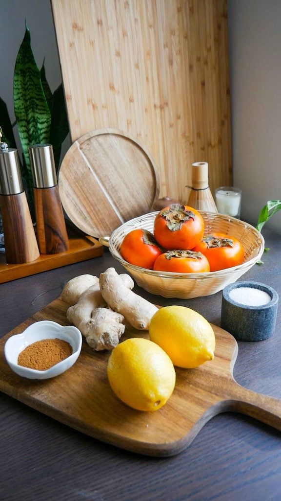 A collection of baking ingredients including whole Fuyu persimmons, sticks of butter, and small bowls of sugar and cinnamon.