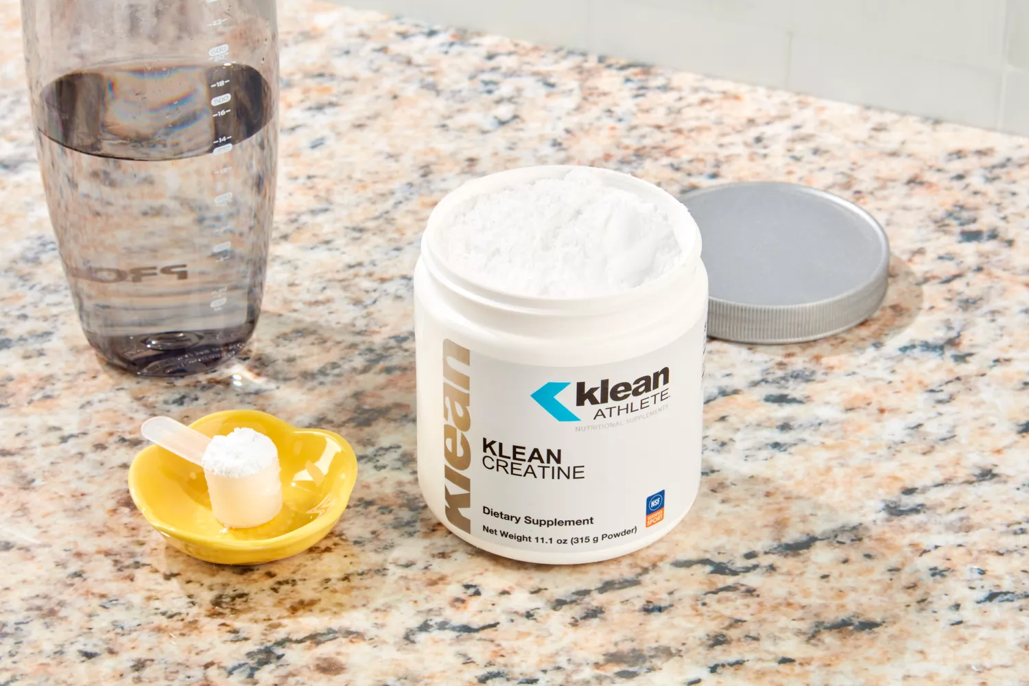 An open container of Klean Athlete Creatine powder on a granite countertop with a measuring scoop.