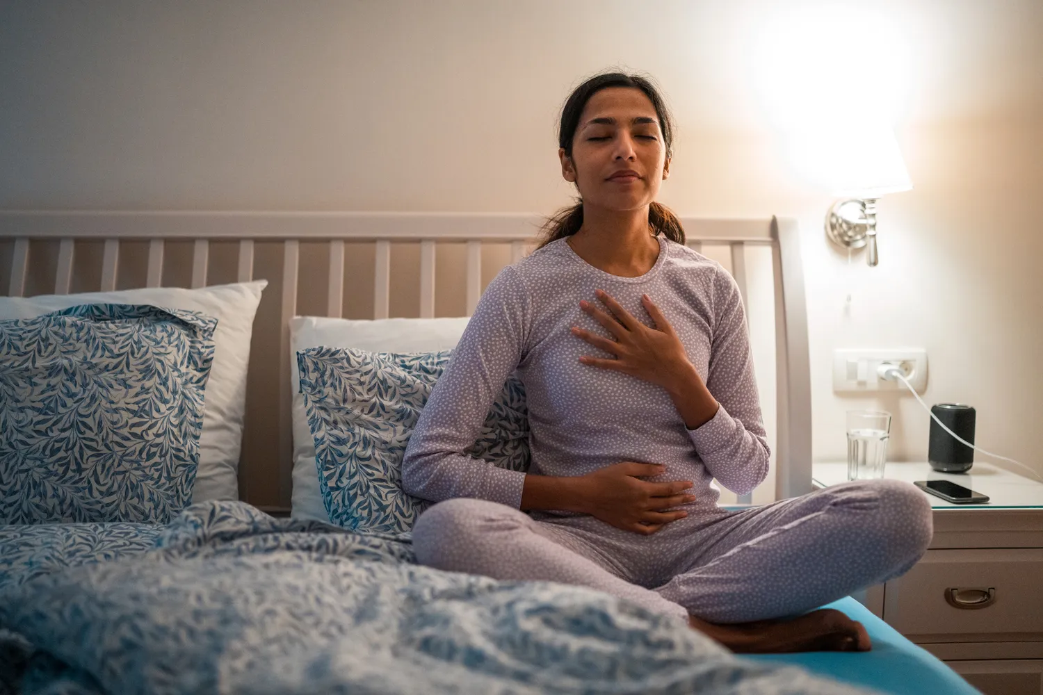 A person sitting cross-legged in pajamas on a bed, meditating before sleep.