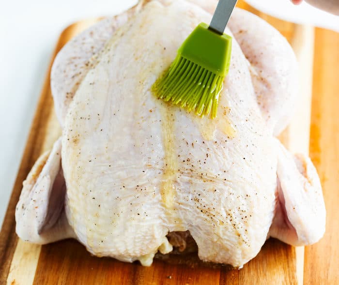 A person using a green pastry brush to spread melted butter over a raw whole chicken.