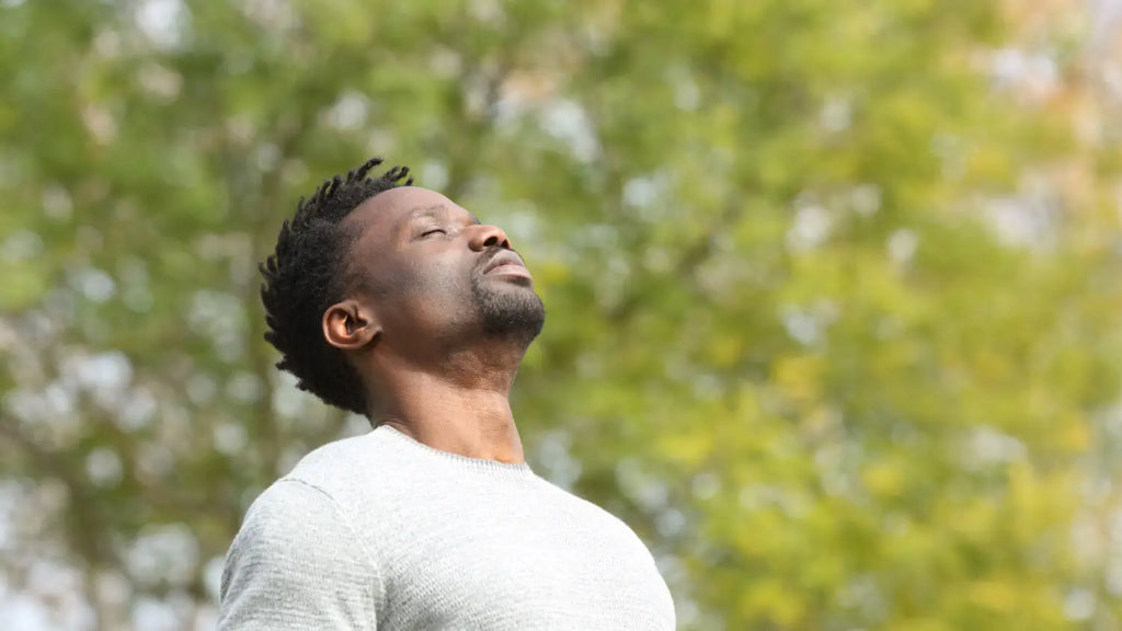 A man standing in a lush green natural setting with his eyes closed, practicing deep breathing.