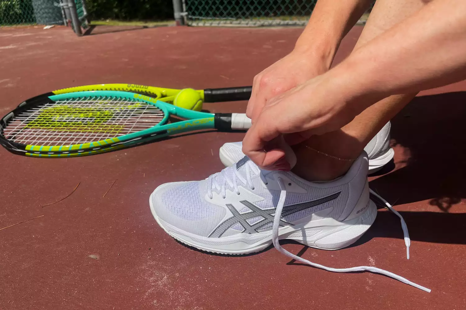 A person tying the laces of a white Asics tennis shoe on a court.