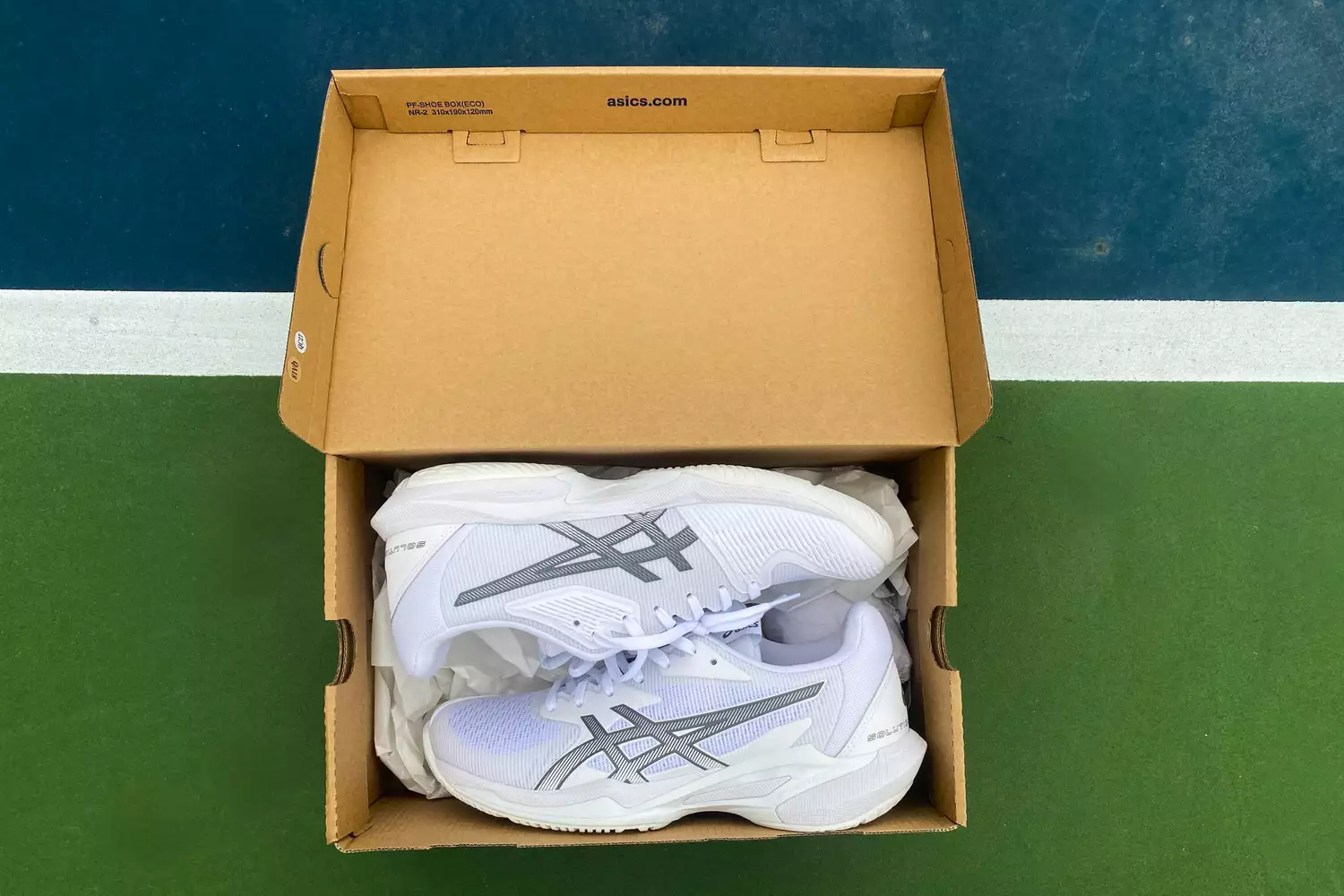 A pair of white ASICS Solution Speed FF 3 tennis shoes inside their original box on a tennis court.